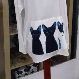 Biała bluzka z długim rękawem i nadrukiem trzech czarnych kotów na kieszeni, prezentowana na manekinie na tle sklepowych półek.