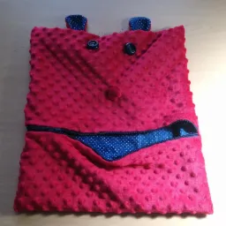 Różowa poduszka z wypustkami, imitująca twarz z guzikami jako oczami, małym pomponem jako nosem i rozpinanym zamkiem błyskawicznym ukazującym niebieską podszewkę w białe kropki jako ustami.