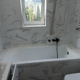 Nowoczesna łazienka z białą wanną, czarną armaturą i płytkami imitującymi marmur, okno z widokiem na zewnątrz, w trakcie wykańczania.