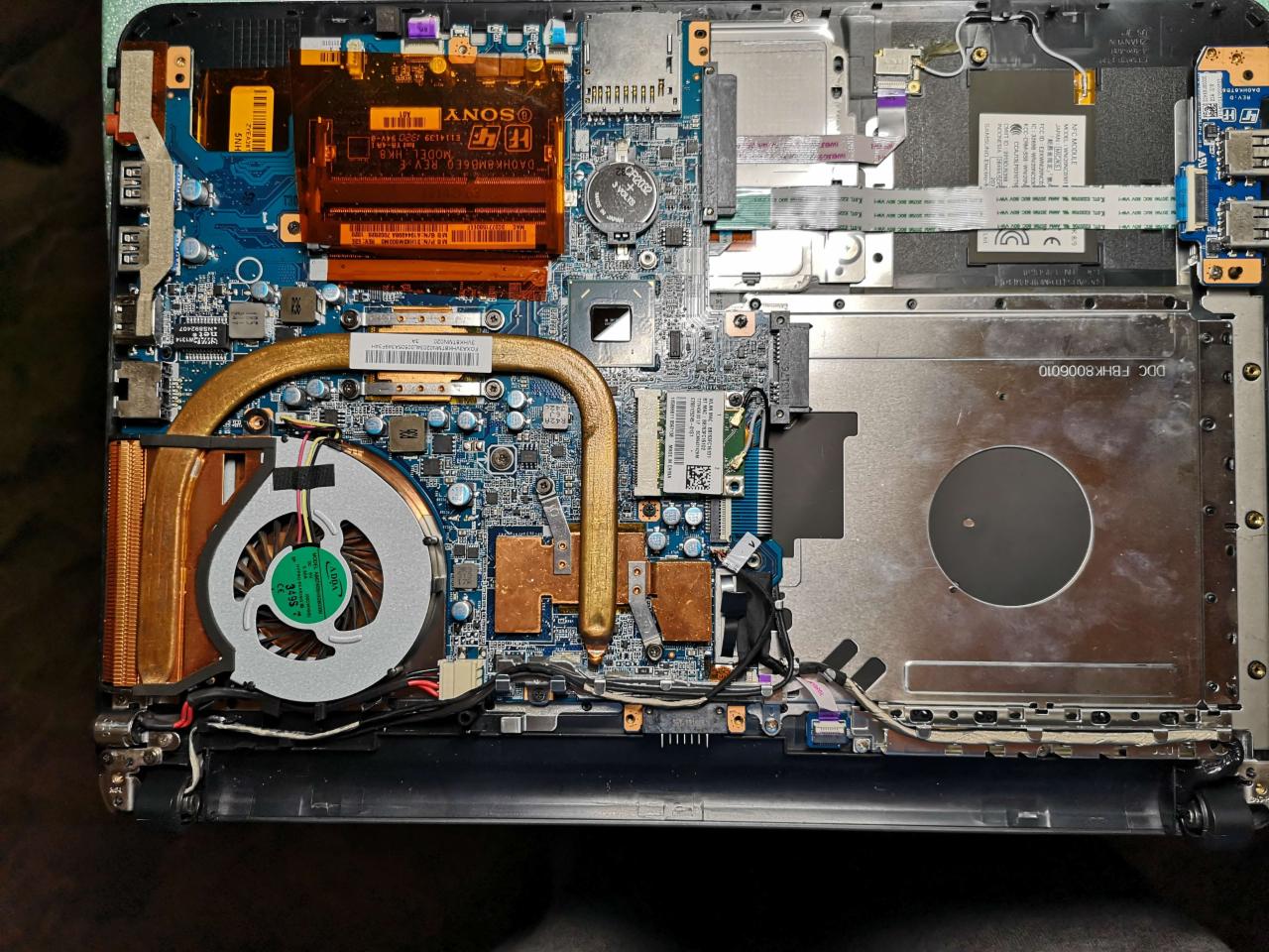 Wnętrze otwartego laptopa z widoczną płytą główną, wentylatorem, radiatorem, baterią CMOS i innymi komponentami. Widoczne ślady użytkowania i drobne zabrudzenia.