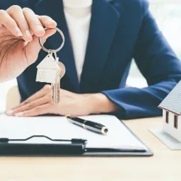 Osoba w granatowym garniturze trzyma klucze do domu, model domu, umowa i długopis na biurku.
