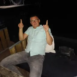 Mężczyzna w okularach siedzi na drewnianej ławce na zewnątrz, wskazując palcami do góry, w tle ciemność i konstrukcja sceniczna.