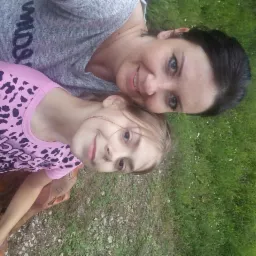 Portret uśmiechniętej kobiety i dziewczynki, leżących na trawie w słoneczny dzień, z widocznym fragmentem rzeźby z drewna.