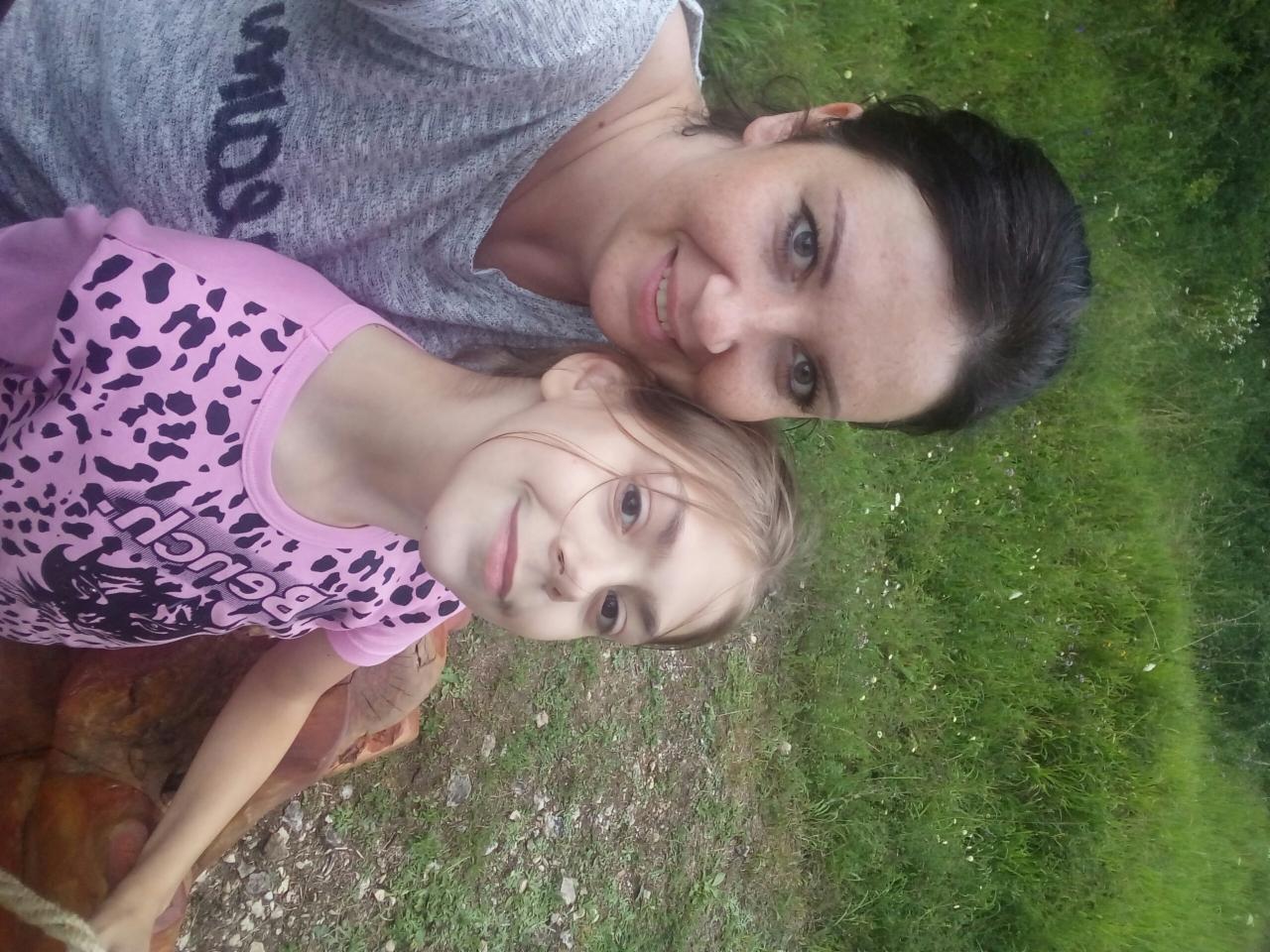 Portret uśmiechniętej kobiety i dziewczynki, leżących na trawie w słoneczny dzień, z widocznym fragmentem rzeźby z drewna.