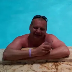 Mężczyzna w okularach na głowie, zanurzony w basenie, opiera się o kamienną krawędź, trzymając szklankę w dłoni. Błękitna woda wypełnia kadr.