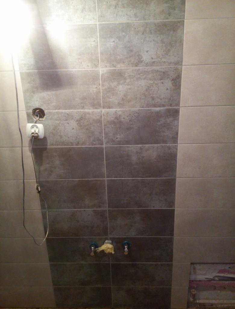 Ściana łazienki z częściowo ułożonymi szarymi i beżowymi płytkami, widoczne wystające kable elektryczne z gniazdkiem i rurki wodne zabezpieczone pianką montażową, ukazujące etap prac wykończeniowych.