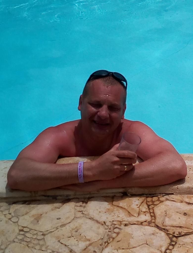 Mężczyzna w okularach na głowie, zanurzony w basenie, opiera się o kamienną krawędź, trzymając szklankę w dłoni. Błękitna woda wypełnia kadr.