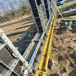 Sylwester Marczyński - Szczegółowy widok szalunków z betonu na placu budowy, z żółtymi elementami wzmacniającymi i widocznym samochodem w tle, w słoneczny dzień.