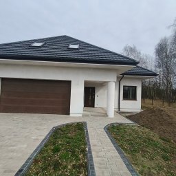 Sylwester Marczyński - Parterowy dom z brązową bramą garażową i ciemną dachówką. Jasna elewacja, kostka brukowa i trawnik przed budynkiem. Widoczne drzewa w tle.