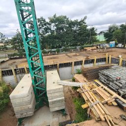 Sylwester Marczyński - Fundamenty budynku w trakcie budowy z widocznym dźwigiem, szalunkami i zbrojeniami. Ujęcie z góry, ukazujące etap prac ziemnych i przygotowania do wylewania betonu.