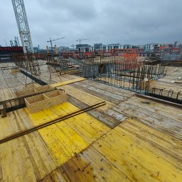 Sylwester Marczyński - Żółte szalunki stropowe z widocznym zbrojeniem na budowie wielopiętrowego budynku w pochmurny dzień. W tle dźwigi i budynki.