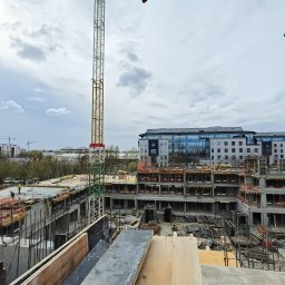Sylwester Marczyński - Budowa budynku wielorodzinnego w trakcie realizacji. Widoczne szalunki, zbrojenia, dźwigi i pracujący robotnicy w żółtych kamizelkach. W tle inne budynki.