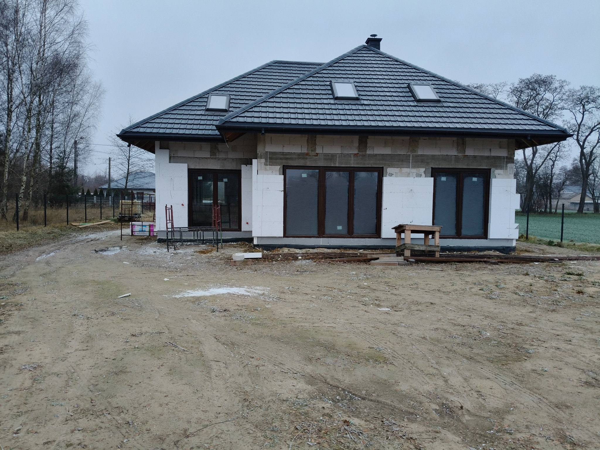 Surowy stan domu murowanego z oknami i ciemną dachówką, widoczne pustaki i ocieplenie styropianem, teren budowy w pochmurny dzień.