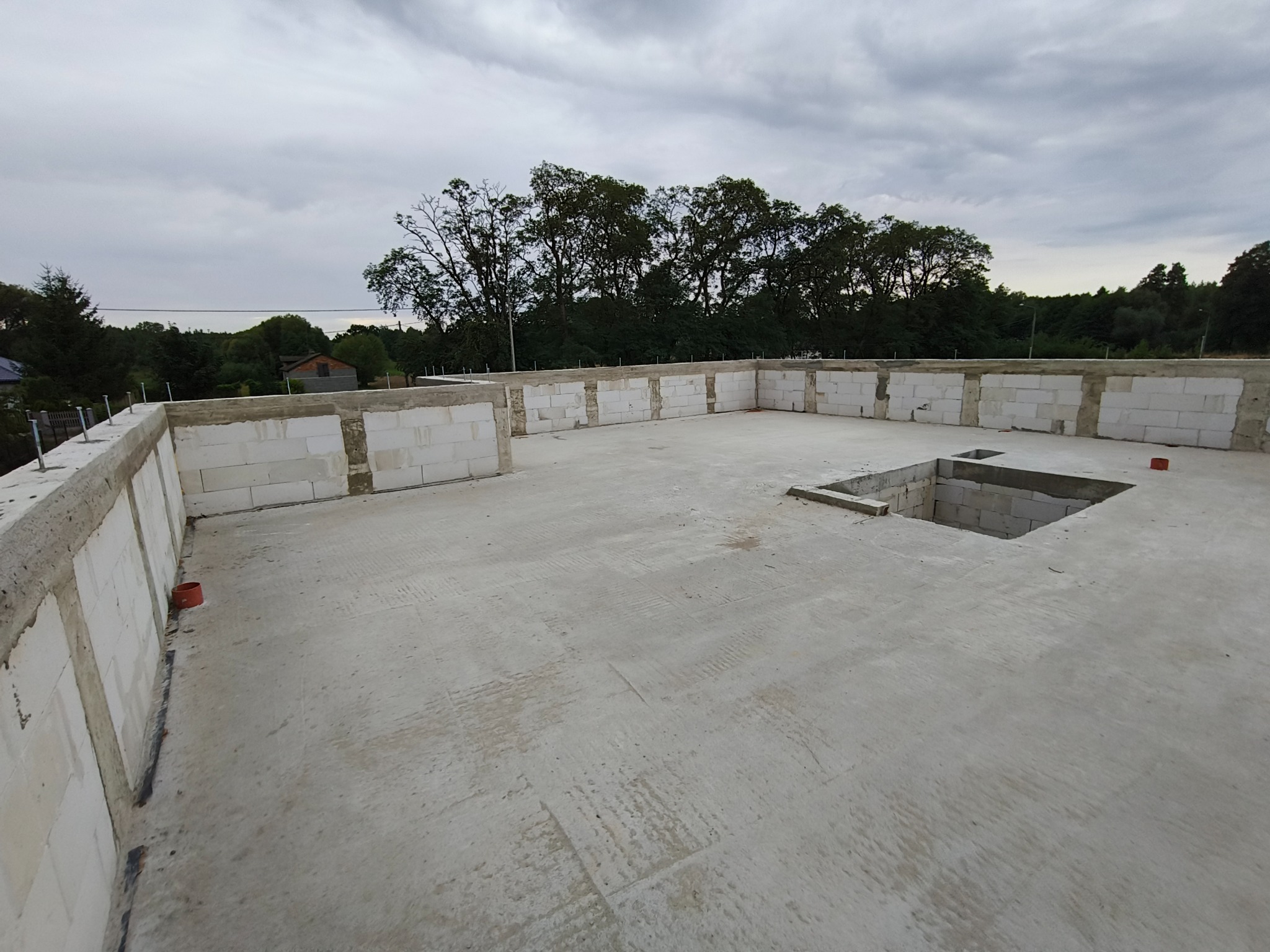 Niedokończony strop budynku z betonu komórkowego, widoczne ślady po szalunkach, w tle zieleń drzew i pochmurne niebo.