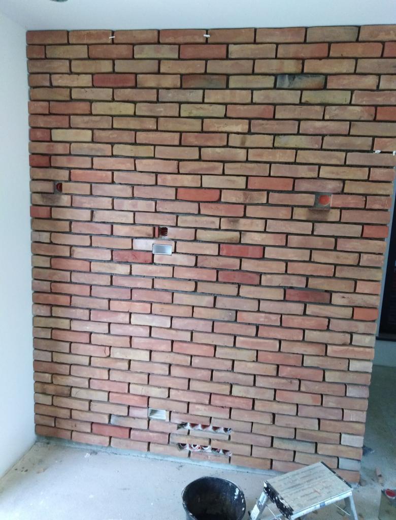 Ściana z cegieł klinkierowych w trakcie montażu, widoczne otwory na instalacje, wiadro i drabinka malarska w dolnej części kadru.