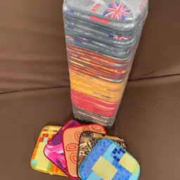 Stos zapakowanych w folię, kolorowych podkładek pod krzesła, niektóre z motywem flagi brytyjskiej i abstrakcyjnymi wzorami, leżące na brązowym tle.