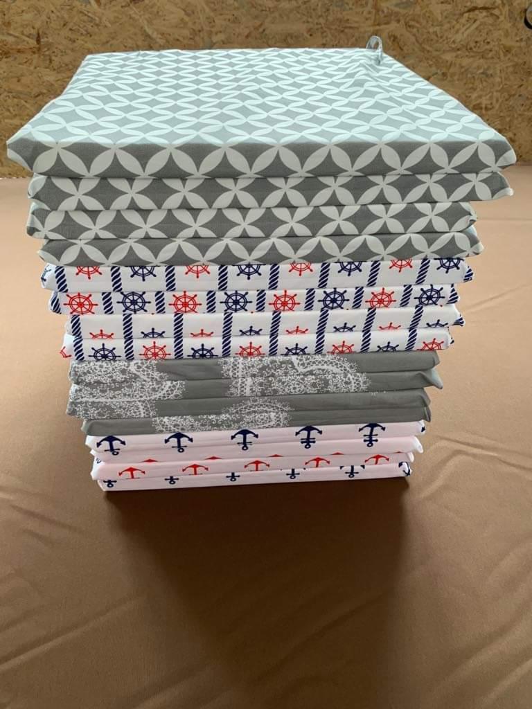 Stos złożonych, świeżo uszytych poduszek dekoracyjnych z różnymi wzorami marynistycznymi i geometrycznymi na brązowym tle.