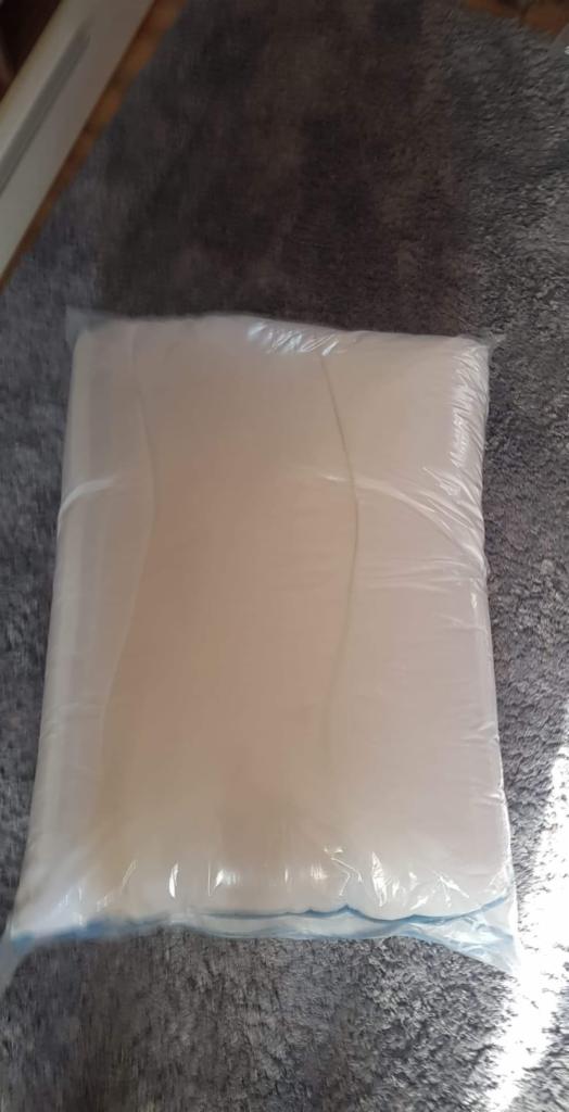 Duża, biała poduszka w plastikowym opakowaniu, leżąca na szarym, puszystym dywanie.
