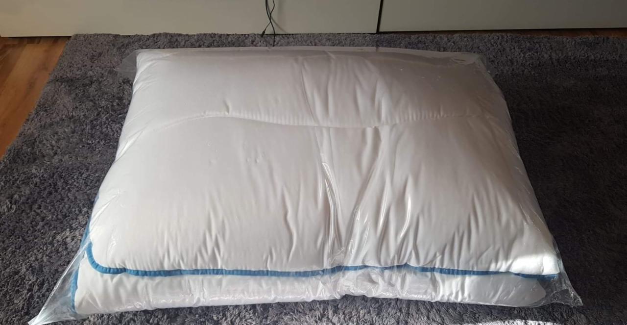 Nowa, biała poduszka z niebieskim obszyciem, zapakowana w przezroczysty plastikowy worek, leżąca na szarym dywanie.