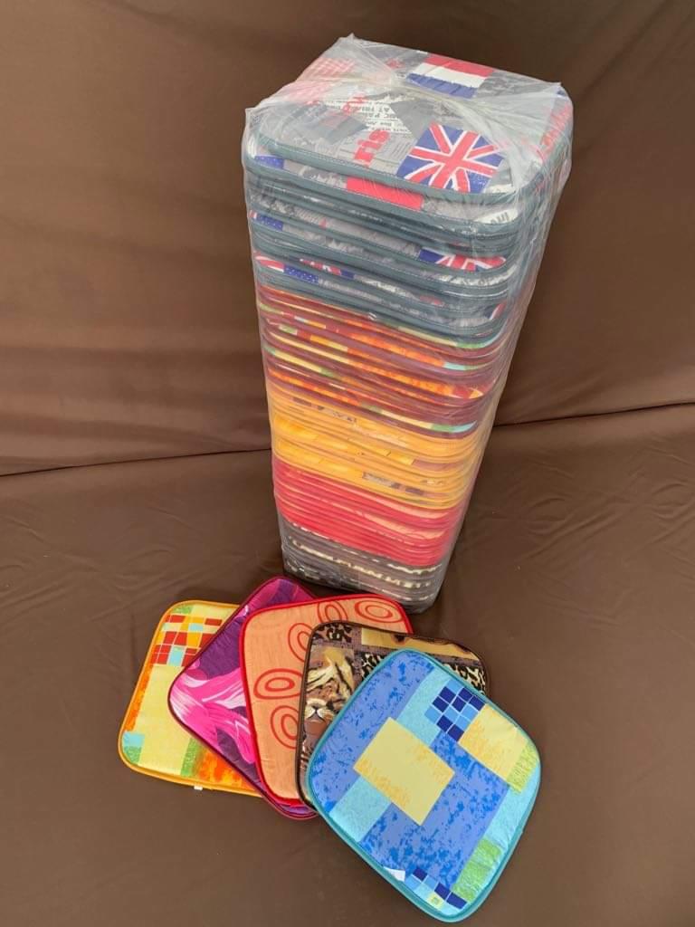 Stos zapakowanych w folię, kolorowych podkładek pod krzesła, niektóre z motywem flagi brytyjskiej i abstrakcyjnymi wzorami, leżące na brązowym tle.
