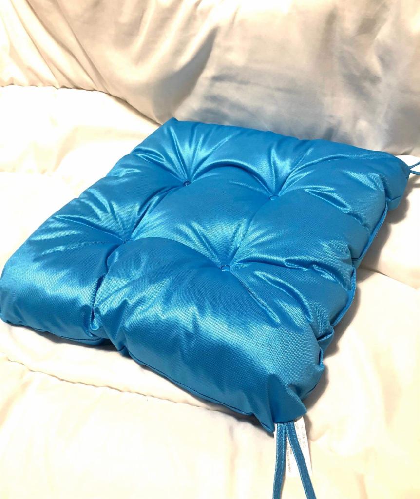 Błękitna, pikowana poduszka na krzesło z połyskującego materiału, leżąca na białej pościeli z widocznymi sznurkami do mocowania.