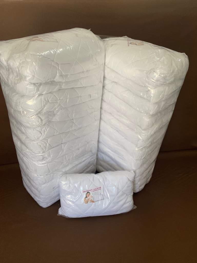 Dwa stosy zapakowanych w folię, białych, pikowanych poduszek na brązowym tle, z mniejszą poduszką z naklejką na pierwszym planie.