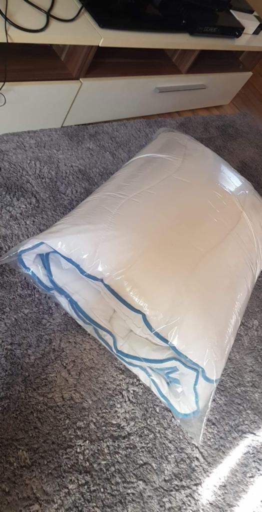 Biała kołdra w przezroczystym opakowaniu z niebieską lamówką leżąca na szarym dywanie shaggy, w tle szafka RTV.