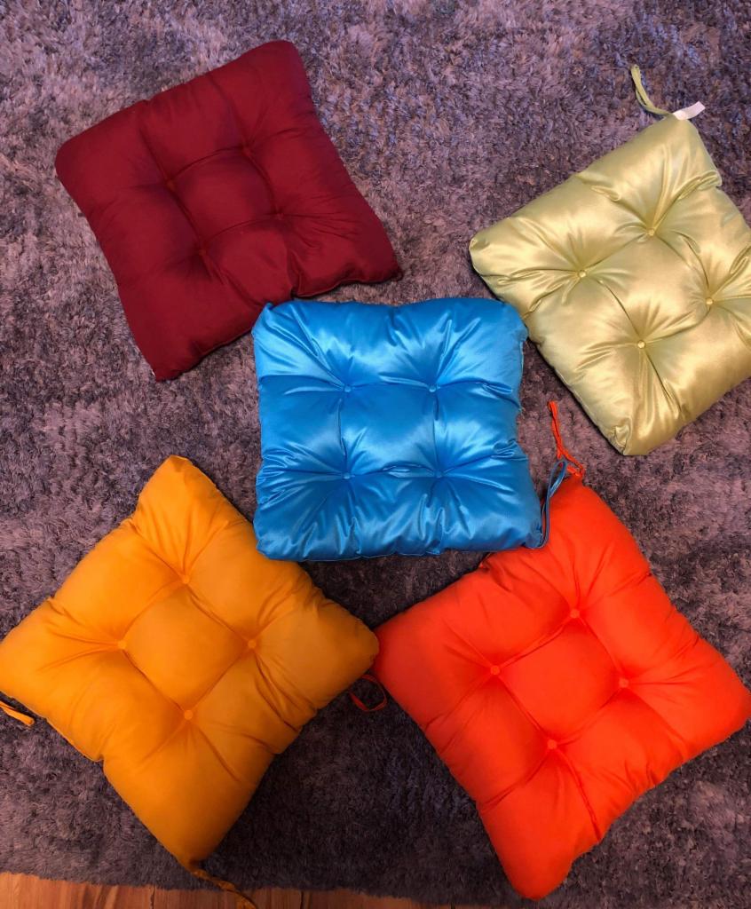 Kolekcja pięciu kwadratowych poduszek siedziskowych w różnych kolorach (czerwony, zielony, niebieski, żółty, pomarańczowy) z pikowaniami, ułożonych na szarym dywanie o długim włosiu.