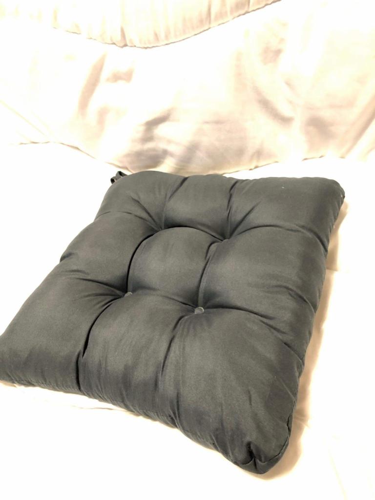 Szara, kwadratowa poduszka na krzesło z guzikami, leżąca na białej pościeli, widziana z góry pod kątem.