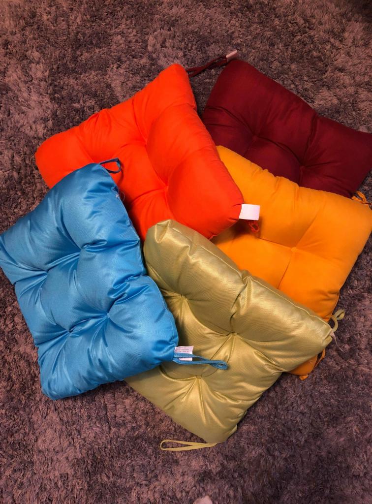 Kaskada kolorowych poduszek na krzesła ogrodowe: turkusowa, limonkowo-zielona, pomarańczowa i bordowa, ułożone na dywanie z długim włosiem.