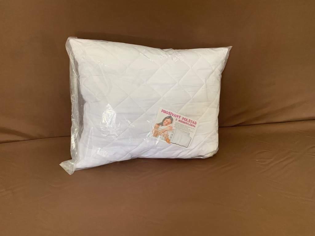 Nowa, biała, pikowana poduszka w przezroczystym opakowaniu foliowym, leżąca na brązowej sofie. Widoczna etykieta produktu w języku obcym.