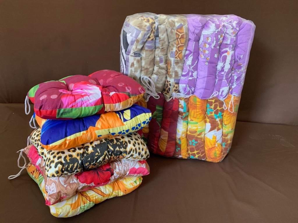 Stos poduszek na krzesła w kwiatowe i zwierzęce wzory, część zapakowana w folię, na brązowej sofie.