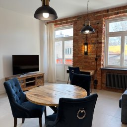 DreamBudTeam Sp.z o. o. - Loftowe wnętrze w Warszawie: ceglana ściana z oknami, drewniany stół otoczony welurowymi krzesłami, lampy w stylu industrialnym, TV na drewnianej komodzie.