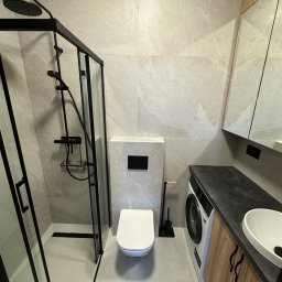 DreamBudTeam Sp.z o. o. - Nowoczesna łazienka z kabiną prysznicową typu walk-in z czarnymi ramami, umywalką nablatową i pralką pod blatem. Minimalistyczny wystrój z szarymi płytkami.