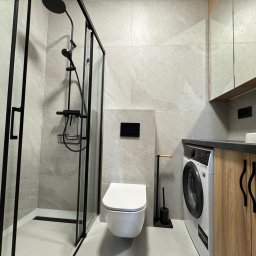 DreamBudTeam Sp.z o. o. - Nowoczesna łazienka z kabiną prysznicową typu walk-in, podwieszaną toaletą i pralką w zabudowie. Szare płytki imitujące kamień i czarne akcenty.