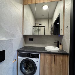 DreamBudTeam Sp.z o. o. - Nowoczesna łazienka: szare płytki, drewniane szafki, pralka pod blatem i lustro z oświetleniem, odbijające kabinę prysznicową. Minimalistyczny design.