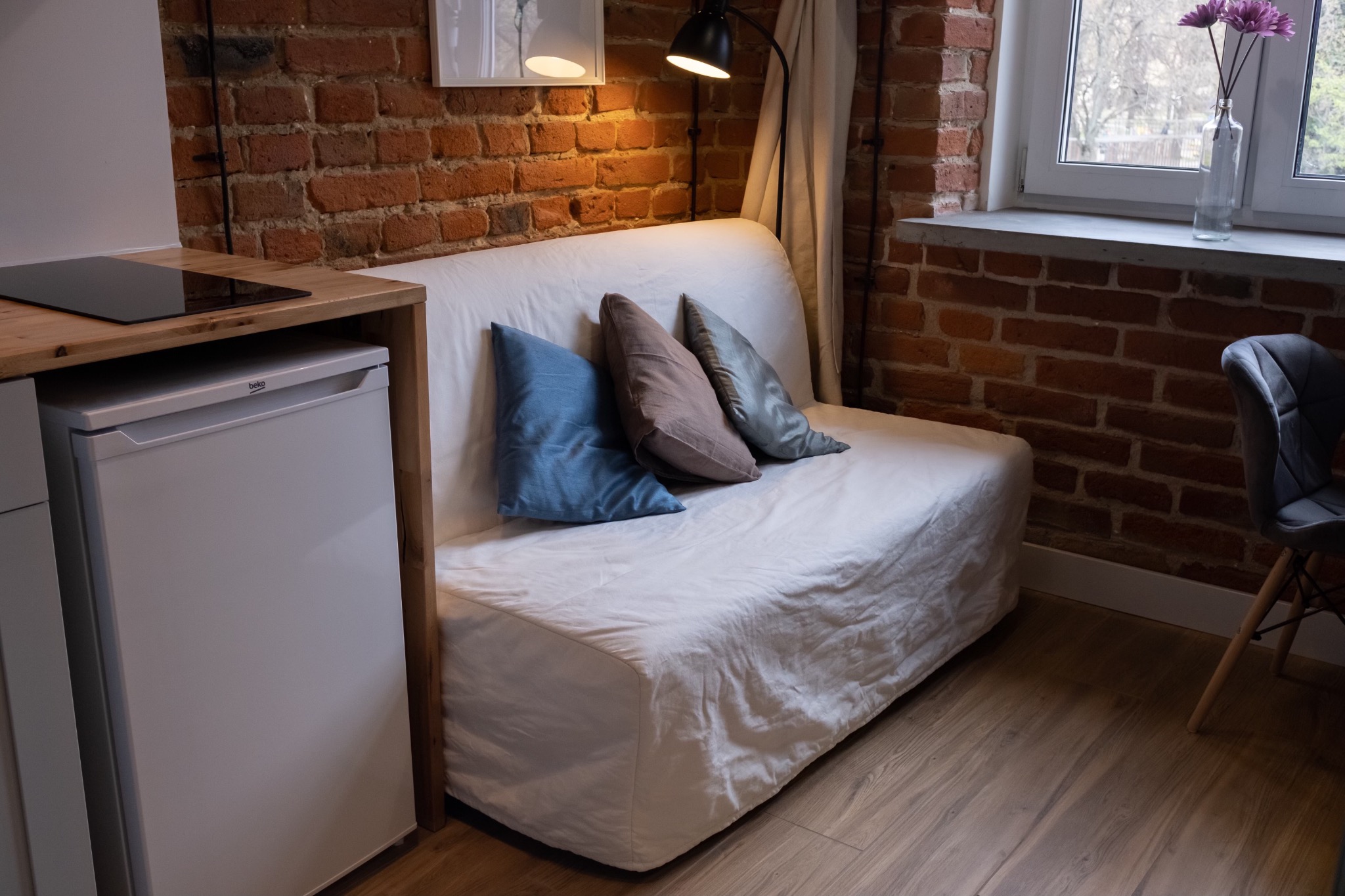 Wnętrze pokoju z odsłoniętą cegłą, białym rozkładanym łóżkiem z trzema poduszkami, aneksem kuchennym i szarym krzesłem na drewnianej podłodze.
