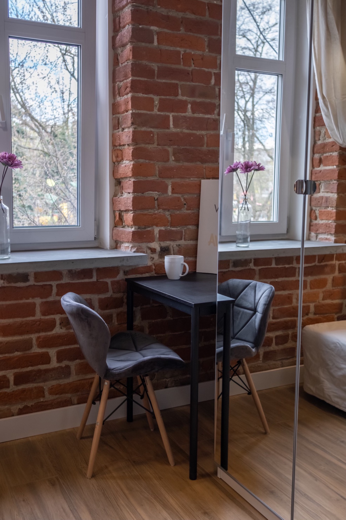 Stylowa aranżacja wnętrza: mały czarny stolik z dwoma szarymi krzesłami na drewnianej podłodze, ceglana ściana z dwoma oknami, wazon z fioletowymi kwiatami i biały kubek na stole.