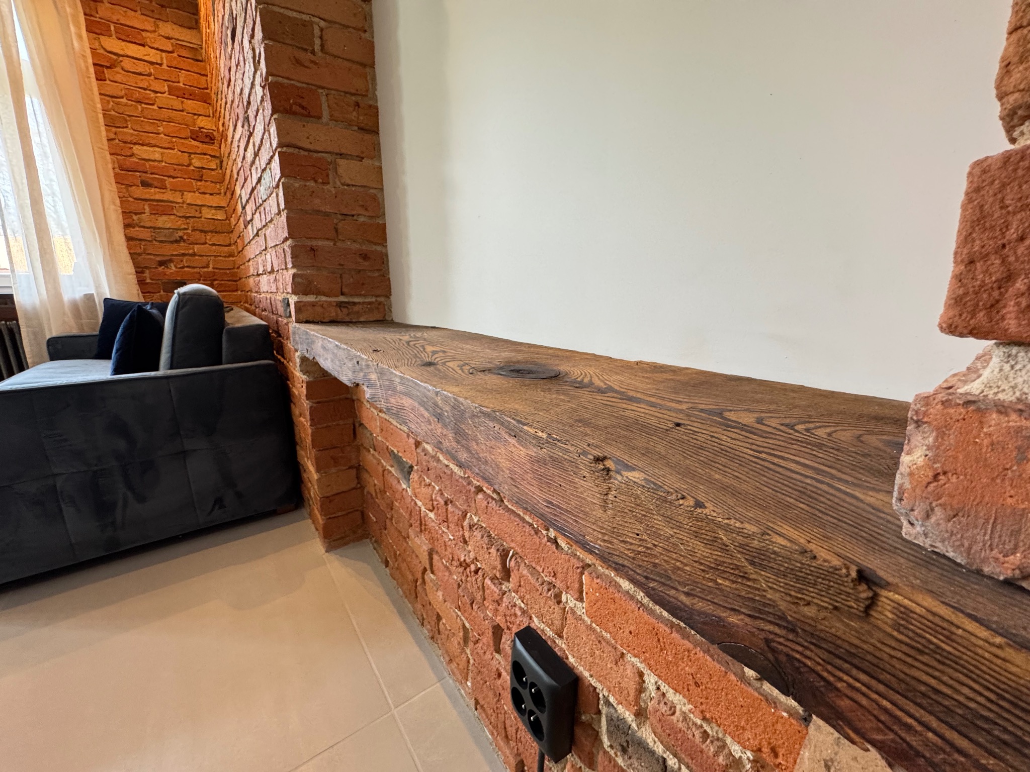 Loftowe wnętrze z ceglaną ścianą, drewnianym blatem i szarą sofą. Widoczne gniazdko elektryczne na ścianie. Naturalne światło z okna.