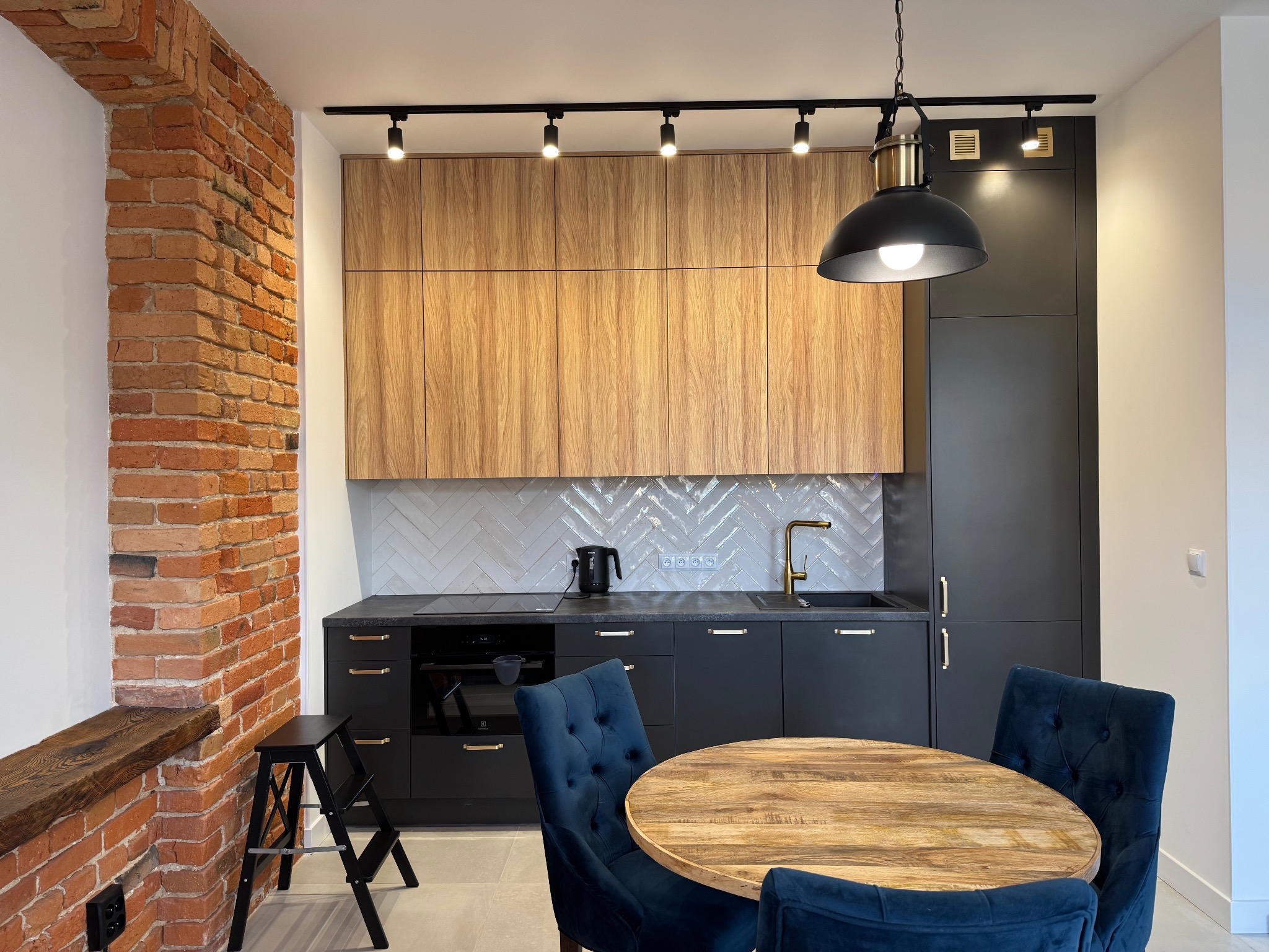 Nowoczesna kuchnia z ceglaną ścianą, drewnianymi szafkami i ciemnymi frontami. Widoczny okrągły stół z krzesłami w kolorze granatowym i oświetlenie punktowe.