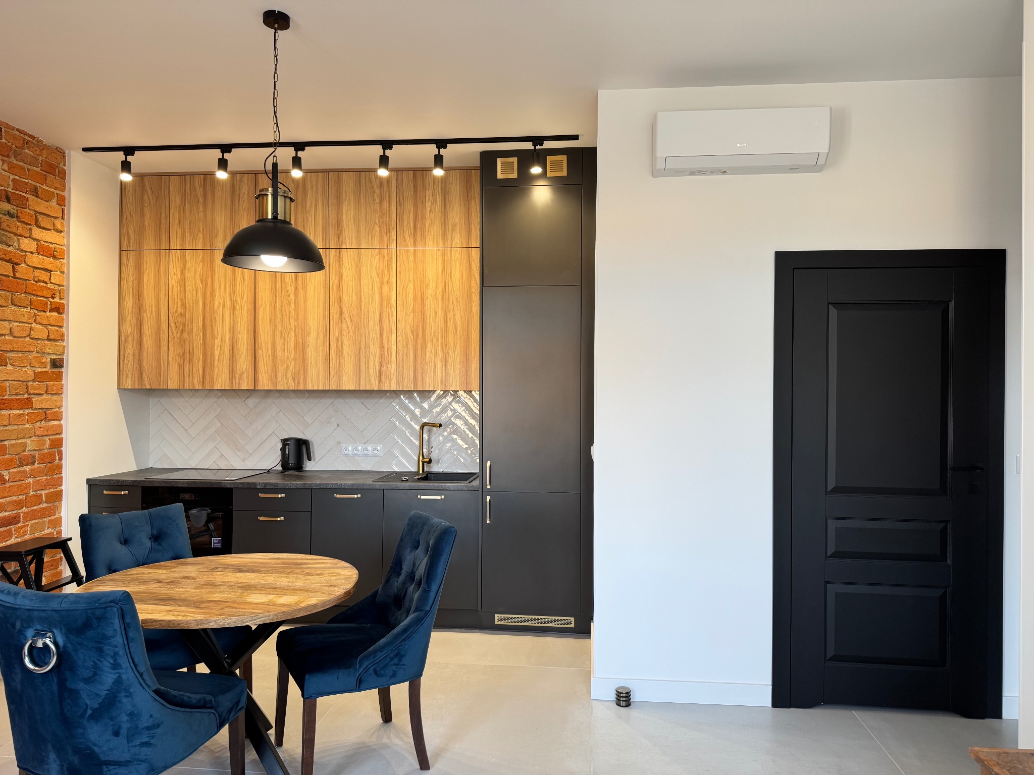 Nowoczesna kuchnia: drewniane szafki, czarne fronty, ceglana ściana, okrągły stół z krzesłami i designerska lampa wisząca. Minimalistyczny design z elementami rustykalnymi.