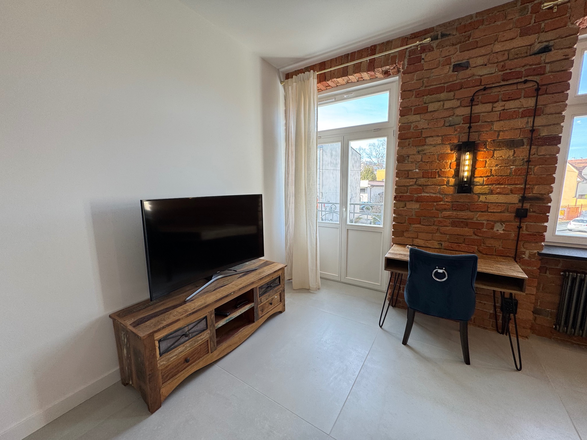 Pokój z ceglaną ścianą, drewnianym biurkiem i szafką RTV. Białe ściany kontrastują z cegłą. Nowoczesne oświetlenie i jasna podłoga dodają elegancji wnętrzu.