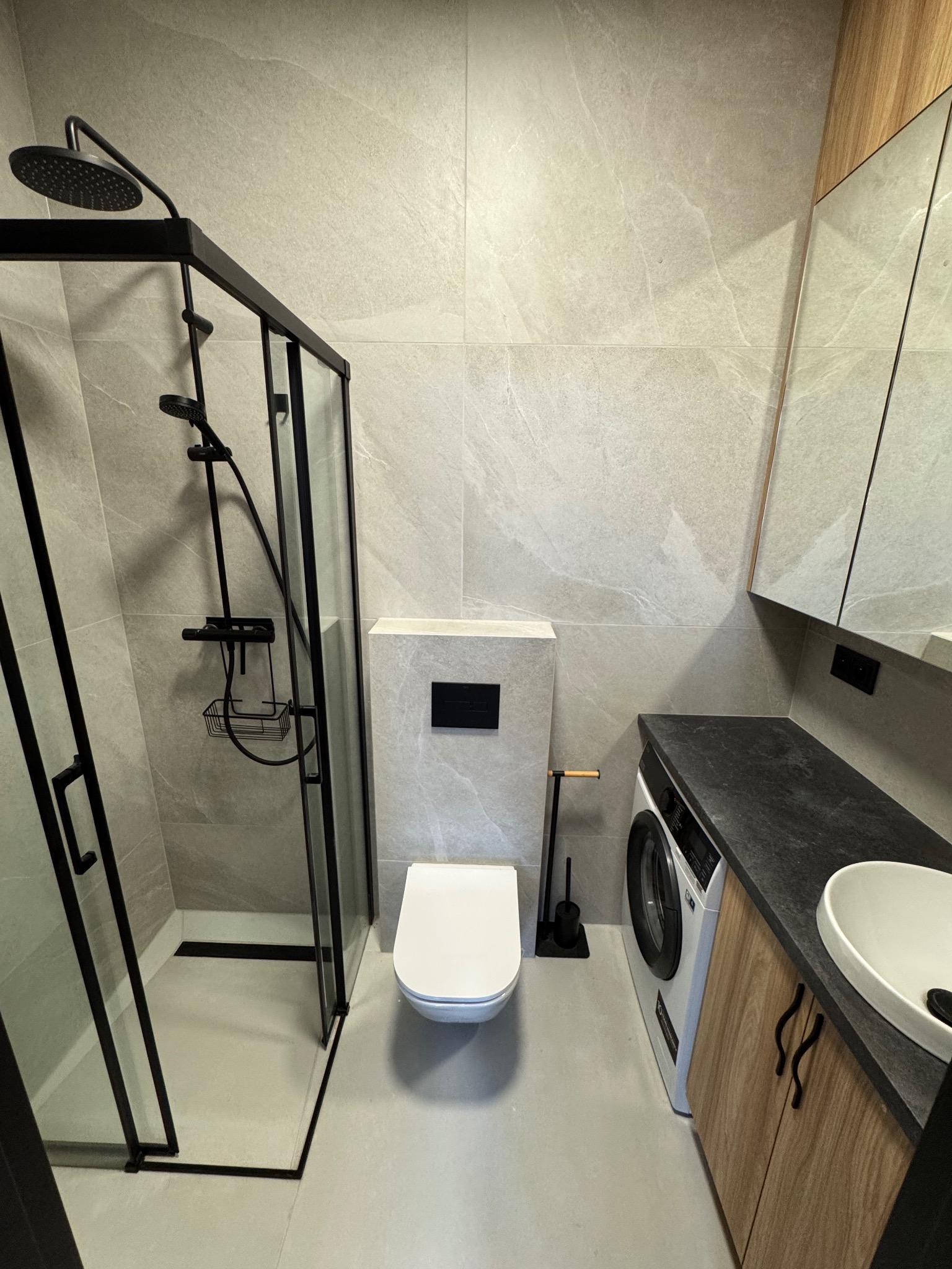 Nowoczesna łazienka z kabiną prysznicową typu walk-in z czarnymi ramami, umywalką nablatową i pralką pod blatem. Minimalistyczny wystrój z szarymi płytkami.