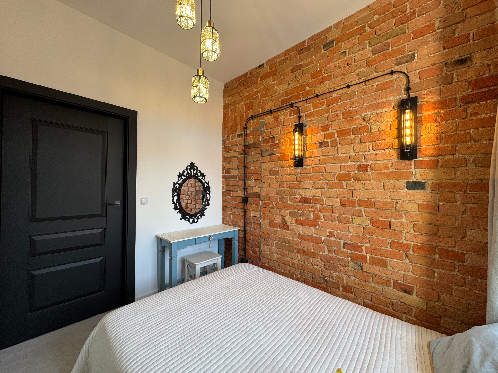 Sypialnia z ceglaną ścianą, oświetleniem industrialnym i czarnymi drzwiami. Widoczny fragment łóżka z białą narzutą i toaletka z lustrem.