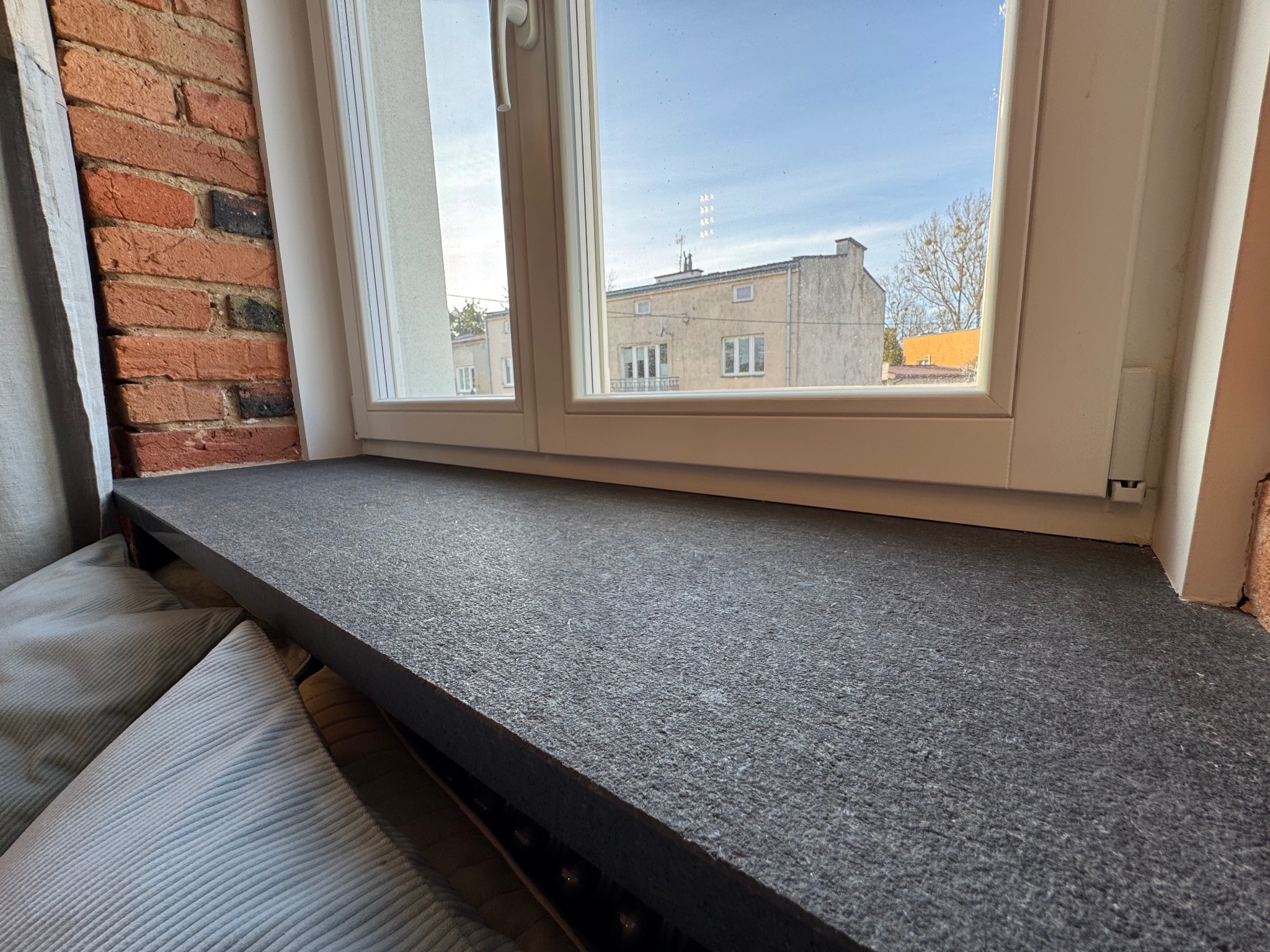 Szary, gruby parapet z widokiem na budynki przez okno, obok ceglana ściana i poduszki. Wykończenie wnętrza w stylu loftowym.