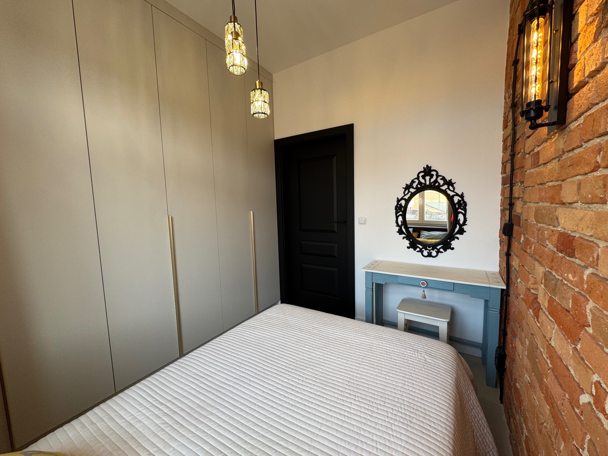 Sypialnia w stylu loft: ceglana ściana z lampą, toaletka z lustrem w ozdobnej ramie, minimalistyczna szafa i wiszące lampy. Biała pościel na łóżku.