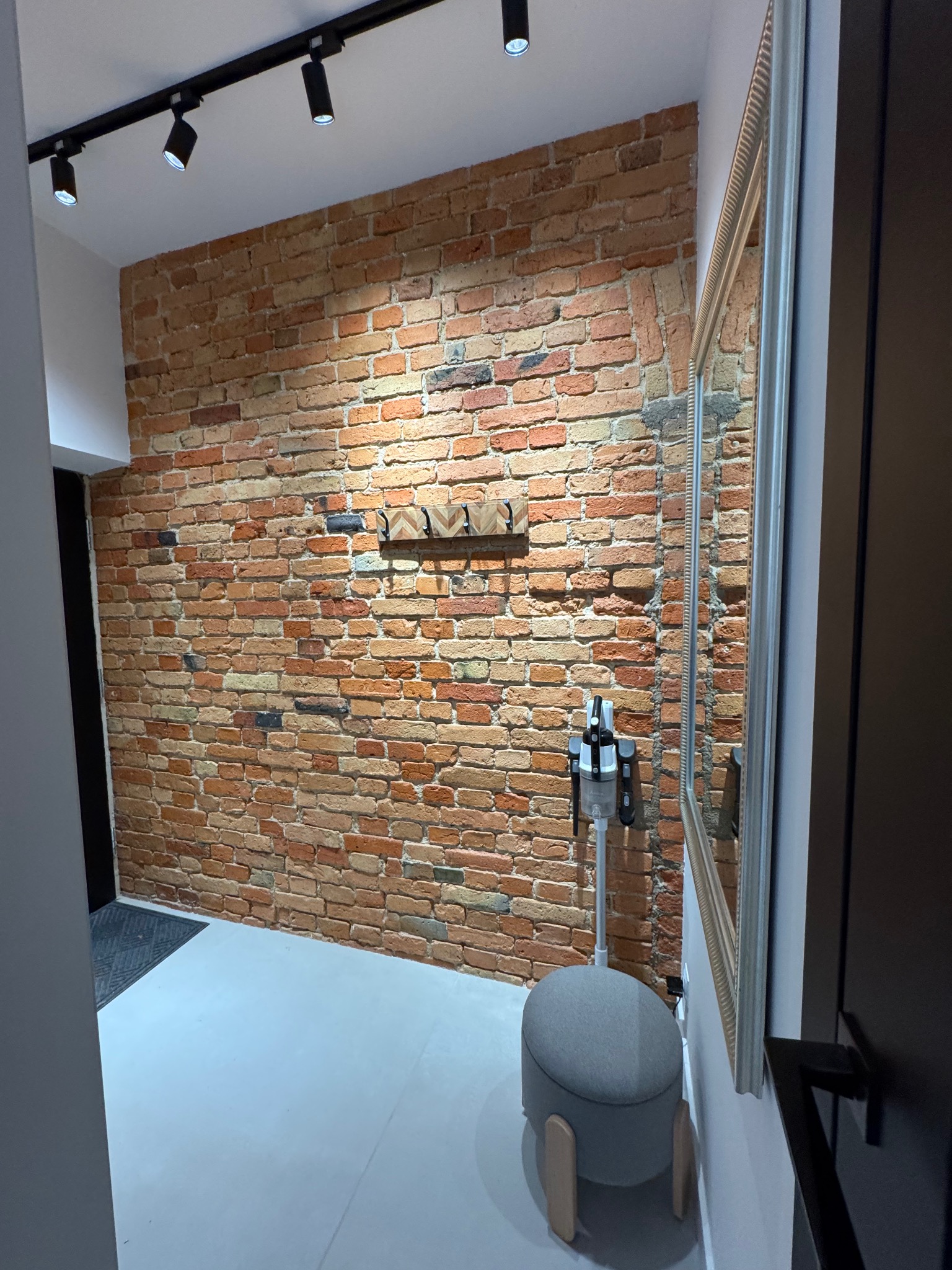 Ceglana ściana z wieszakami, oświetleniem punktowym i lustrem w przedpokoju. W tle widoczny odkurzacz i pufa. Nowoczesne wykończenie wnętrza.