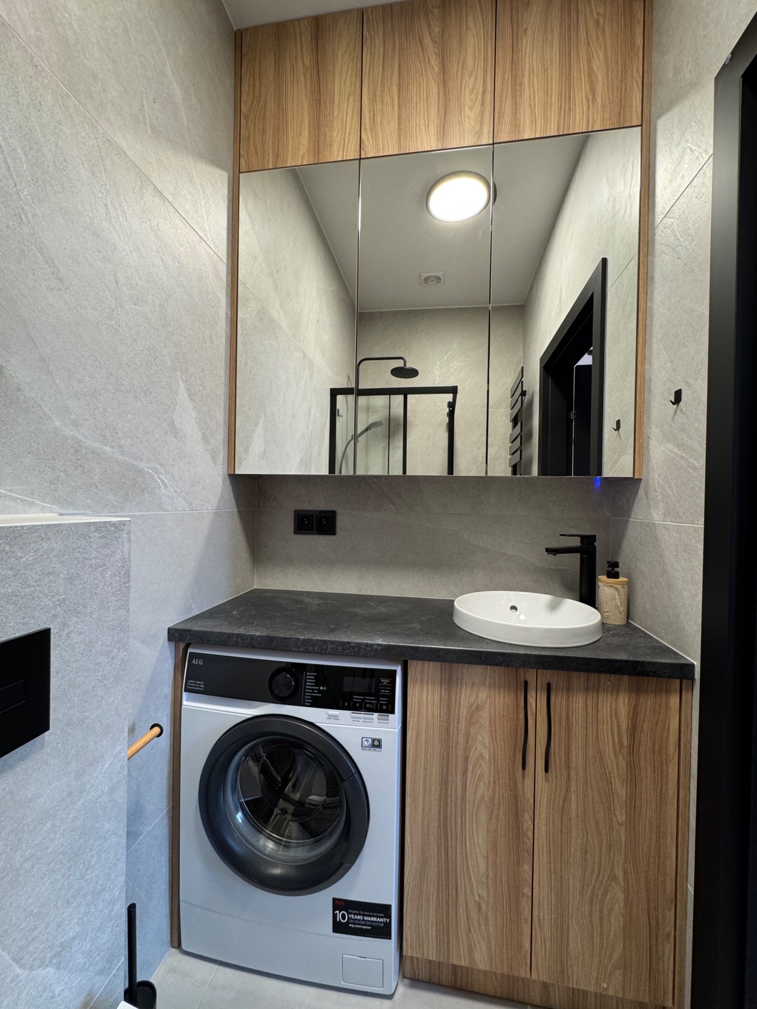 Nowoczesna łazienka: szare płytki, drewniane szafki, pralka pod blatem i lustro z oświetleniem, odbijające kabinę prysznicową. Minimalistyczny design.