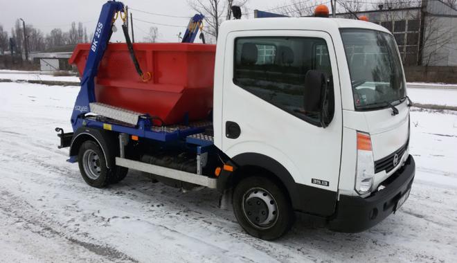 Biała ciężarówka z czerwonym kontenerem na gruz na platformie, stojąca na śniegu w pochmurny dzień.