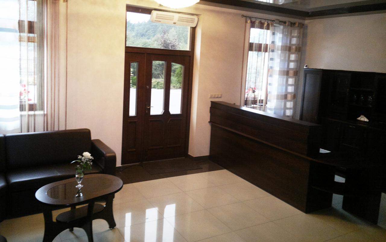 Recepcja hotelowa z ciemnobrązowymi meblami, skórzaną sofą, stolikiem z kwiatami i widokiem na zieleń przez przeszklone drzwi.